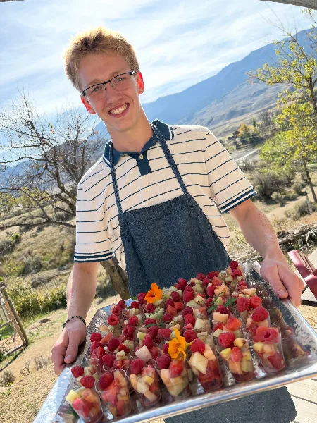 A person wearing a denim apron holds a silver tray filled with small cups of fruit salad garnished with edible flowers, set against a mountainous outdoor background.