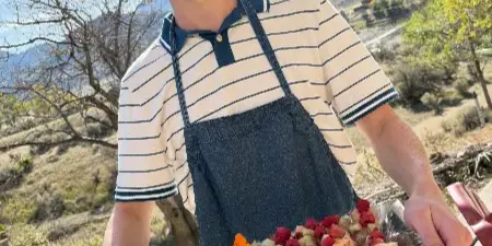 A person wearing a denim apron holds a silver tray filled with small cups of fruit salad garnished with edible flowers, set against a mountainous outdoor background.