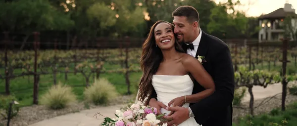 A smiling bride and groom embracing in a vineyard setting, holding a bouquet of flowers.