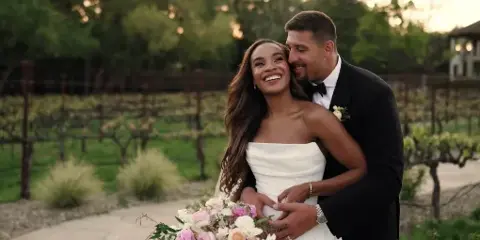 A smiling bride and groom embracing in a vineyard setting, holding a bouquet of flowers.