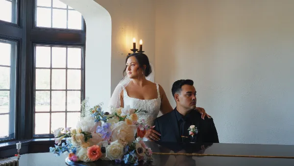 A bride and groom posing together in a bright room with a large floral arrangement in the foreground.