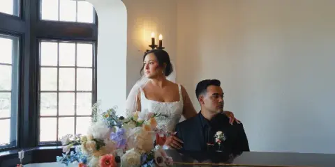 A bride and groom posing together in a bright room with a large floral arrangement in the foreground.