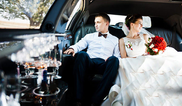 A bride and groom sitting in the back seat of a luxury car with champagne glasses on the table.