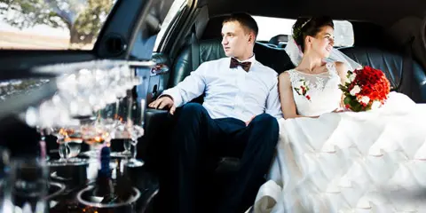 A bride and groom sitting in the back seat of a luxury car with champagne glasses on the table.
