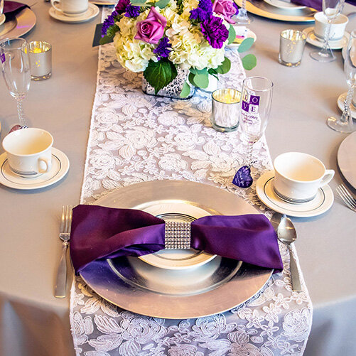 A close-up of a formal table setting featuring a silver charger plate, a purple napkin with a rhinestone ring, and a floral centerpiece.