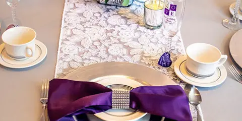A close-up of a formal table setting featuring a silver charger plate, a purple napkin with a rhinestone ring, and a floral centerpiece.