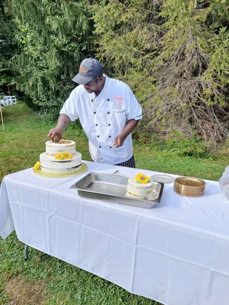 A chef in a white Homestyle Catering uniform preparing a two-tiered cake at an outdoor event.