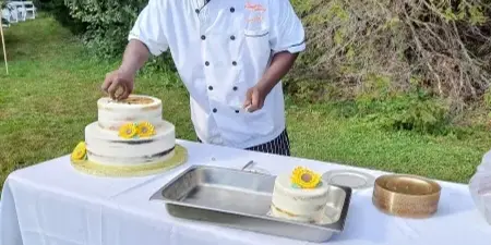 A chef in a white Homestyle Catering uniform preparing a two-tiered cake at an outdoor event.