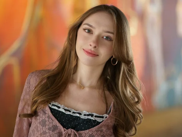 Portrait of a young woman with long hair wearing a pink lace top against a colorful painted backdrop.