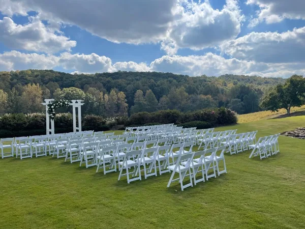Outdoor wedding ceremony setup featuring rows of white folding chairs and a white floral arch on a grassy lawn.
