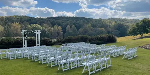 Outdoor wedding ceremony setup featuring rows of white folding chairs and a white floral arch on a grassy lawn.