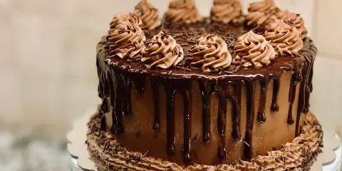 A chocolate celebration cake with dripping ganache and piped frosting swirls on a white cake stand.
