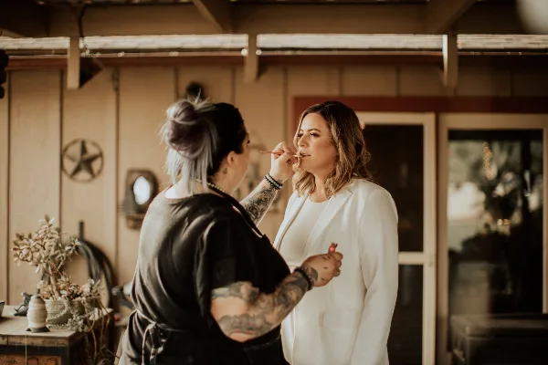 A makeup artist with tattoos applying makeup to a woman in a white blazer.