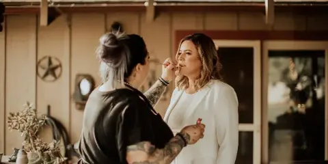 A makeup artist with tattoos applying makeup to a woman in a white blazer.