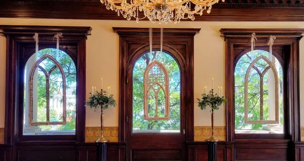 A formal room interior featuring three arched windows, a crystal chandelier, and two brass candlesticks holding floral arrangements.