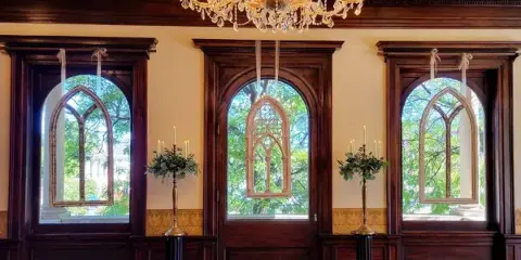 A formal room interior featuring three arched windows, a crystal chandelier, and two brass candlesticks holding floral arrangements.