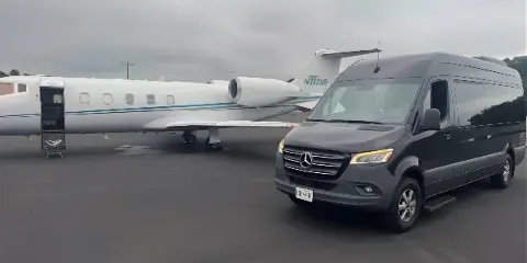 A black Mercedes Sprinter luxury shuttle van parked on an airport tarmac next to a private jet.