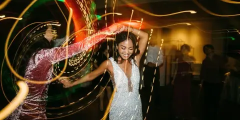 A bride in a white dress dancing joyfully at a wedding reception with light trails in the background.