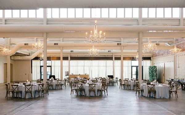 A spacious wedding venue interior with round tables, chandeliers, and a floral green wall installation.