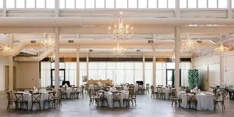 A spacious wedding venue interior with round tables, chandeliers, and a floral green wall installation.