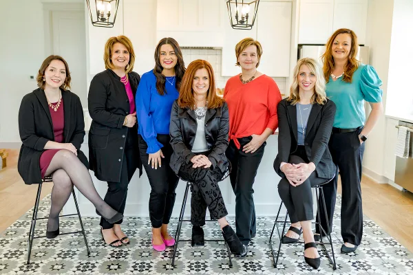 A professional group portrait of seven women posing together in a bright indoor setting.