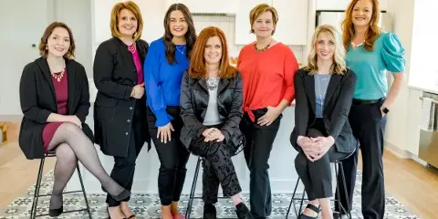 A professional group portrait of seven women posing together in a bright indoor setting.