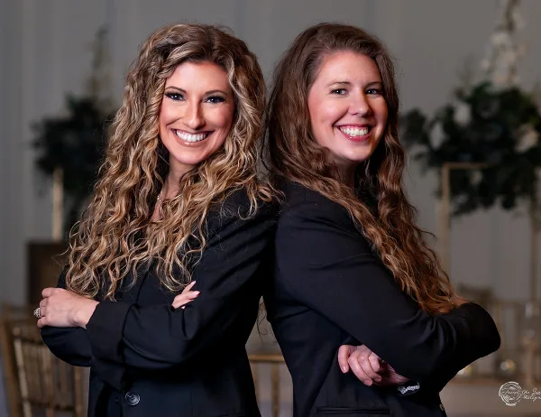 Two smiling women in black blazers standing back-to-back in a decorated event venue.