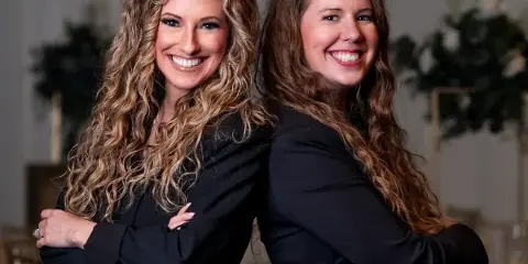 Two smiling women in black blazers standing back-to-back in a decorated event venue.