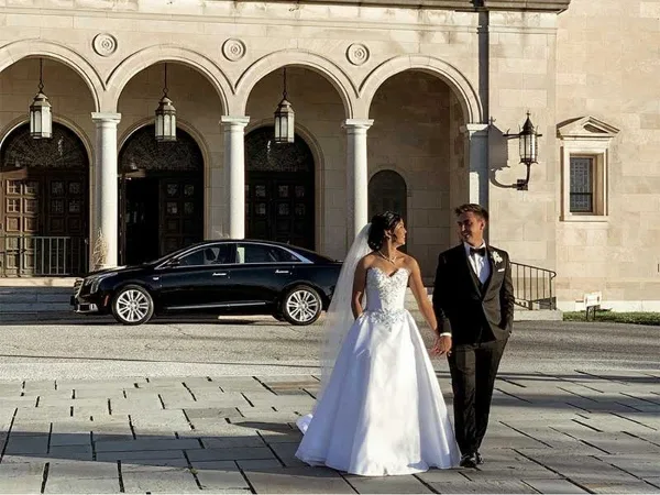 A bride and groom standing in front of a black luxury sedan parked outside a stone building.