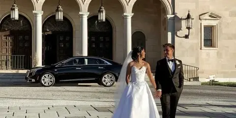 A bride and groom standing in front of a black luxury sedan parked outside a stone building.