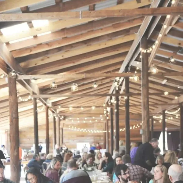 A large group of guests seated at long tables in a rustic wooden venue with string lights.