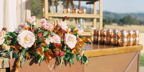 A wooden event bar and shelving unit set up outdoors, featuring a floral arrangement and rows of metallic cups.