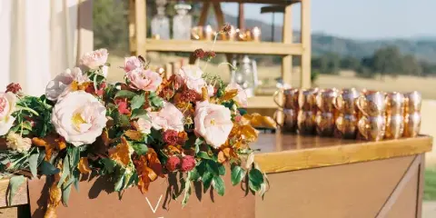 A wooden event bar and shelving unit set up outdoors, featuring a floral arrangement and rows of metallic cups.
