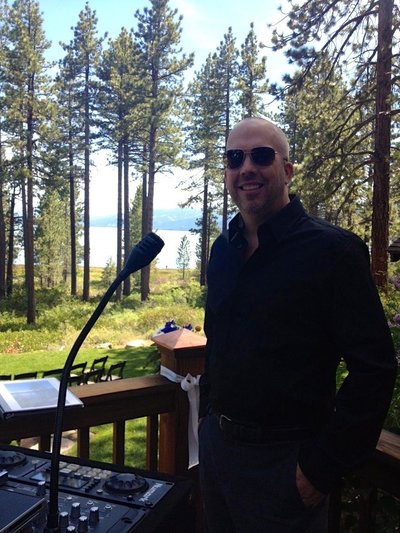 A professional DJ standing next to his audio equipment and microphone at an outdoor venue with a lake view.