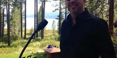 A professional DJ standing next to his audio equipment and microphone at an outdoor venue with a lake view.