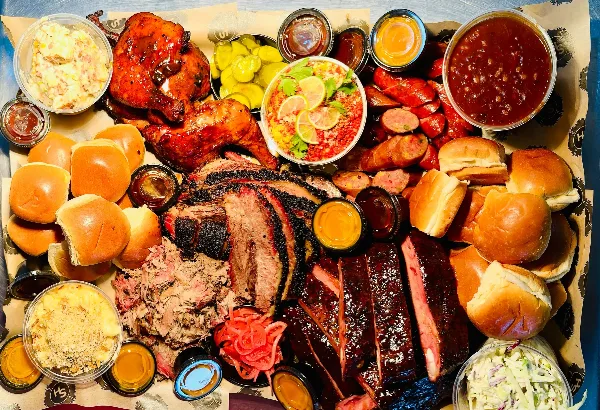 A catering tray filled with smoked brisket, pulled pork, ribs, chicken, buns, and various sides.