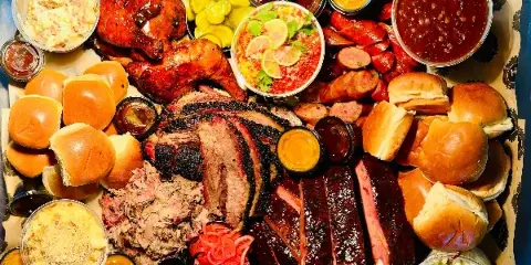 A catering tray filled with smoked brisket, pulled pork, ribs, chicken, buns, and various sides.