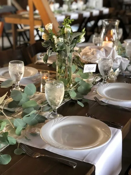 A formal dining table setting featuring white plates, glassware, and a floral centerpiece with eucalyptus and white roses.
