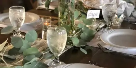 A formal dining table setting featuring white plates, glassware, and a floral centerpiece with eucalyptus and white roses.