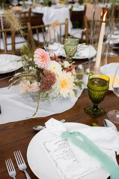 A seasonal floral centerpiece with dahlias and dried grasses on a wooden wedding reception table.