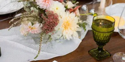 A seasonal floral centerpiece with dahlias and dried grasses on a wooden wedding reception table.