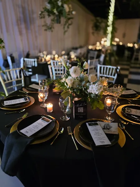 Styled wedding reception table with black linens, gold charger plates, glassware, candles, and a white floral centerpiece in a decorated event space.