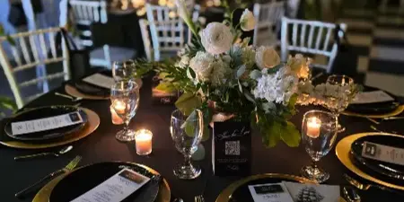Styled wedding reception table with black linens, gold charger plates, glassware, candles, and a white floral centerpiece in a decorated event space.