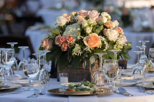 A lush floral centerpiece featuring white and peach roses and greenery in a rustic wooden box, placed on a formally set wedding reception table.