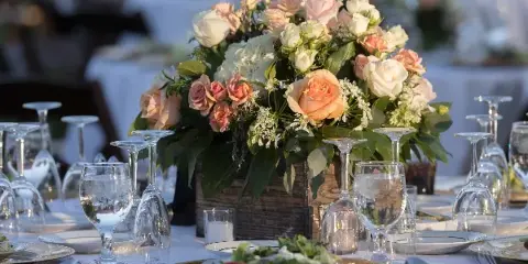 A lush floral centerpiece featuring white and peach roses and greenery in a rustic wooden box, placed on a formally set wedding reception table.
