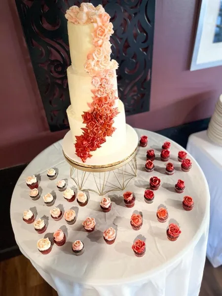 A tiered white wedding cake decorated with a cascading arrangement of pink and red buttercream roses, displayed on a gold stand surrounded by numerous floral cupcakes on a white tablecloth.