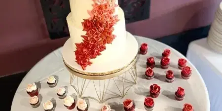 A tiered white wedding cake decorated with a cascading arrangement of pink and red buttercream roses, displayed on a gold stand surrounded by numerous floral cupcakes on a white tablecloth.