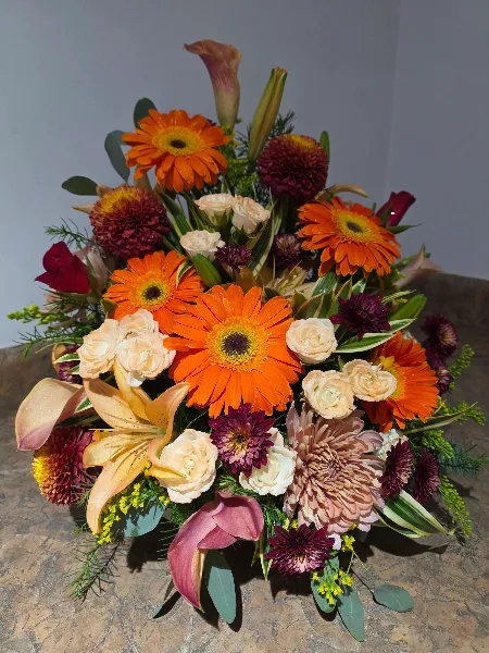 A vibrant floral arrangement featuring orange gerbera daisies, peach lilies, and cream roses on a marble surface.