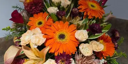A vibrant floral arrangement featuring orange gerbera daisies, peach lilies, and cream roses on a marble surface.