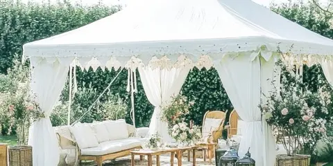 A white event tent set up on a lawn with white draping, a sofa, and floral arrangements.